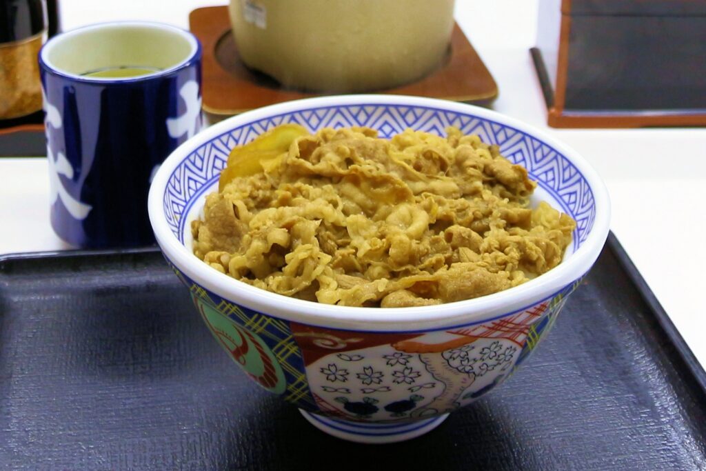 Thin strips of simmered beef and onion covering hot rice in a dark ceramic bowl