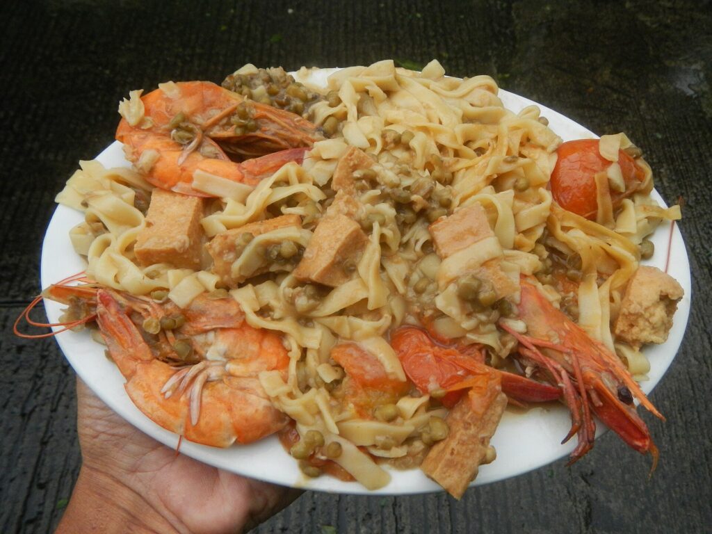 Wide curled egg noodles heaped in a white bowl beside a serving spoon on a counter