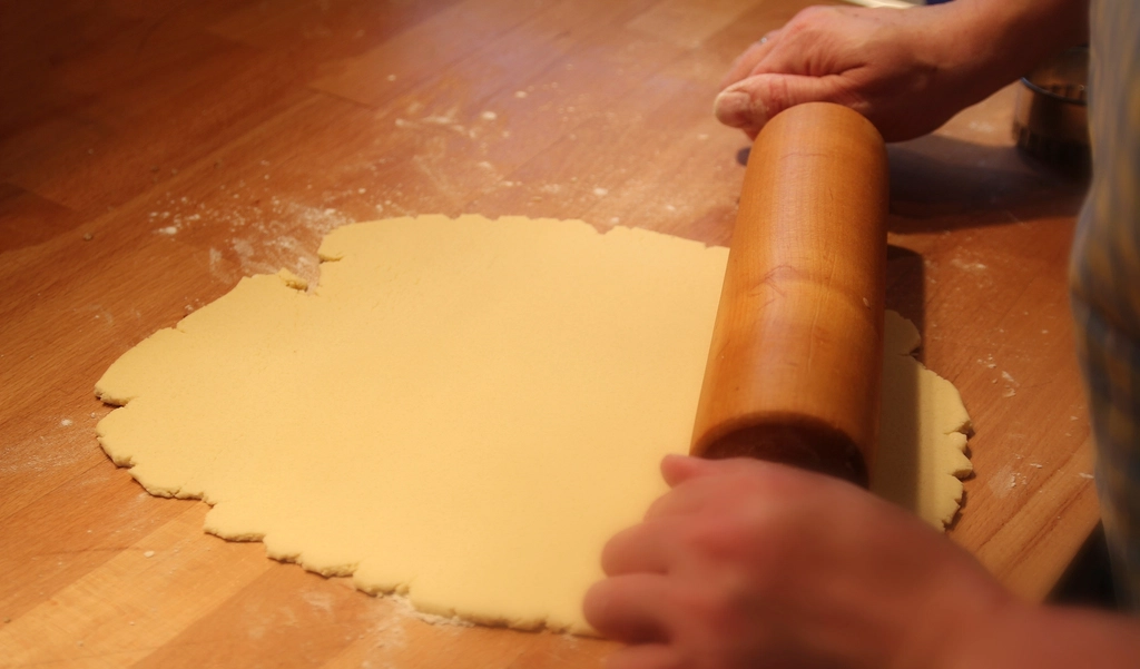 A wooden rolling pin flattening a pale sheet of egg dough on a floured kitchen counter